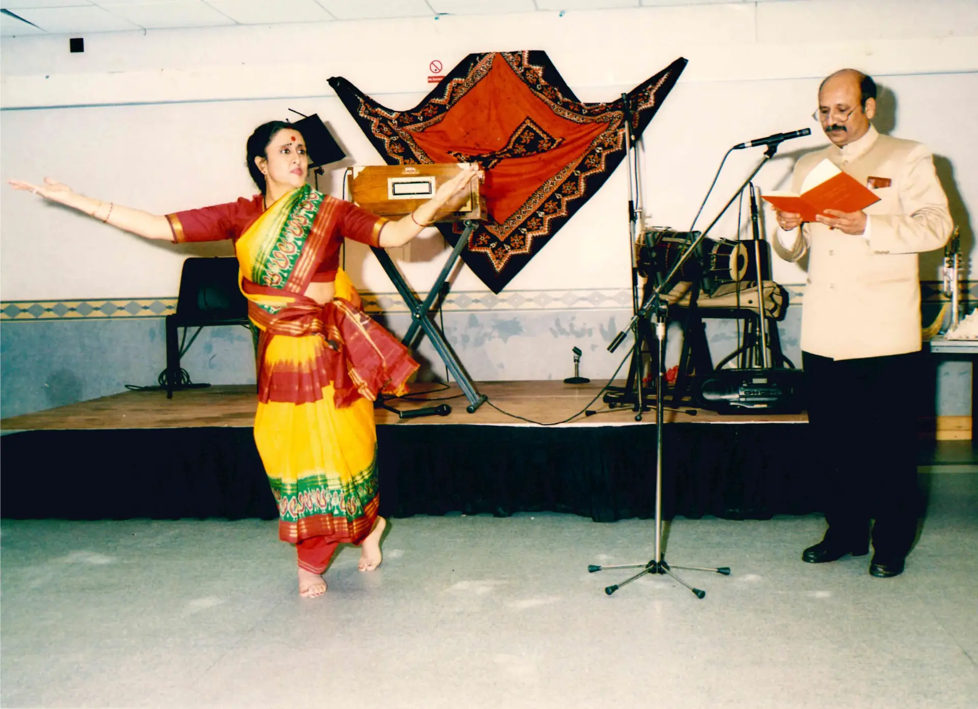 A dancer in a yellow sari performing barefoot on a hall floor while a man in a bandhgala jacket recites from an open book at a microphone